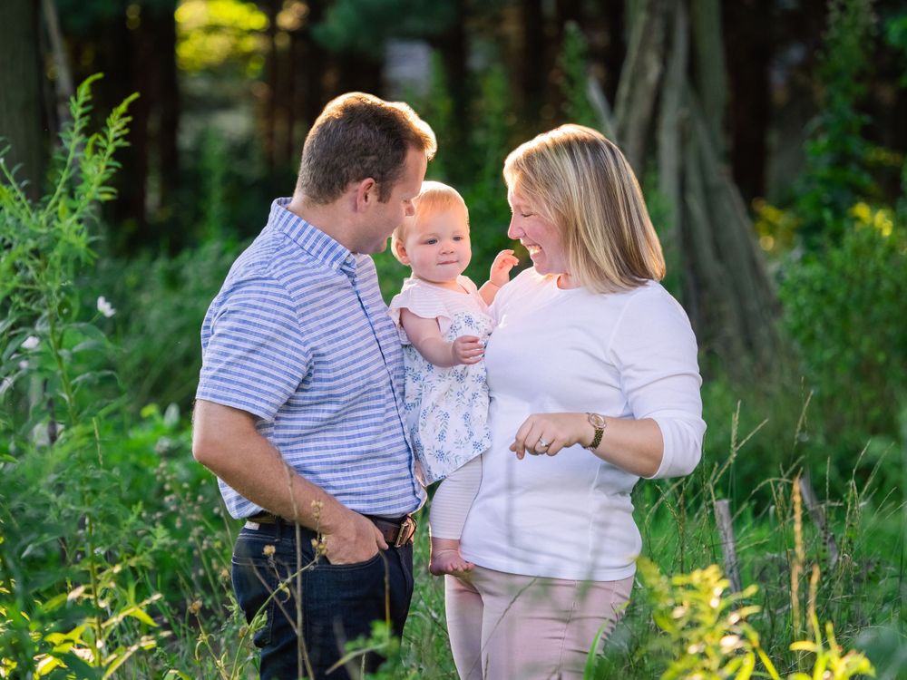 A mother and father smiling at their baby girl in a sunlit meadow during a session for Vermont Family Portraits.