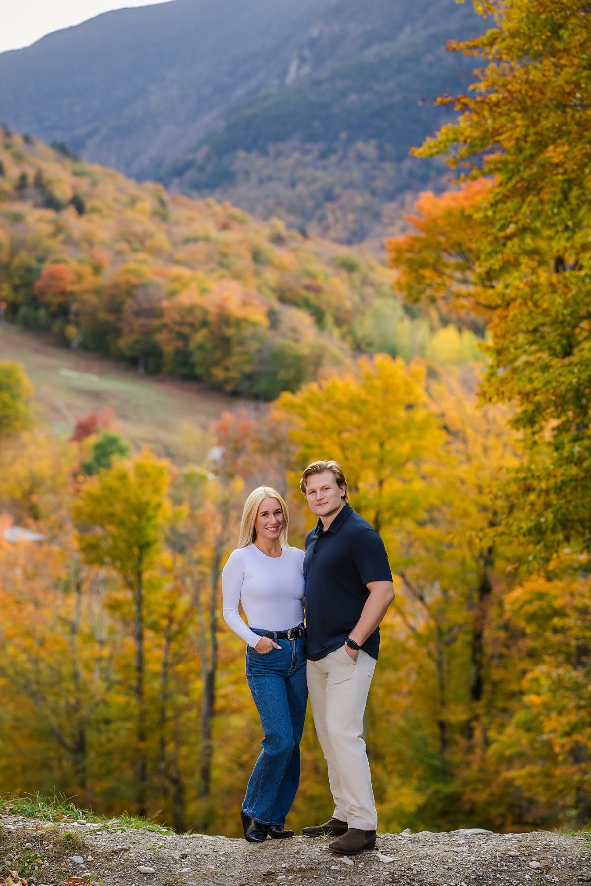 A happy blonde woman and man embracing on a mountain path during peak autumn foliage, captured by a Vermont engagement photographer.